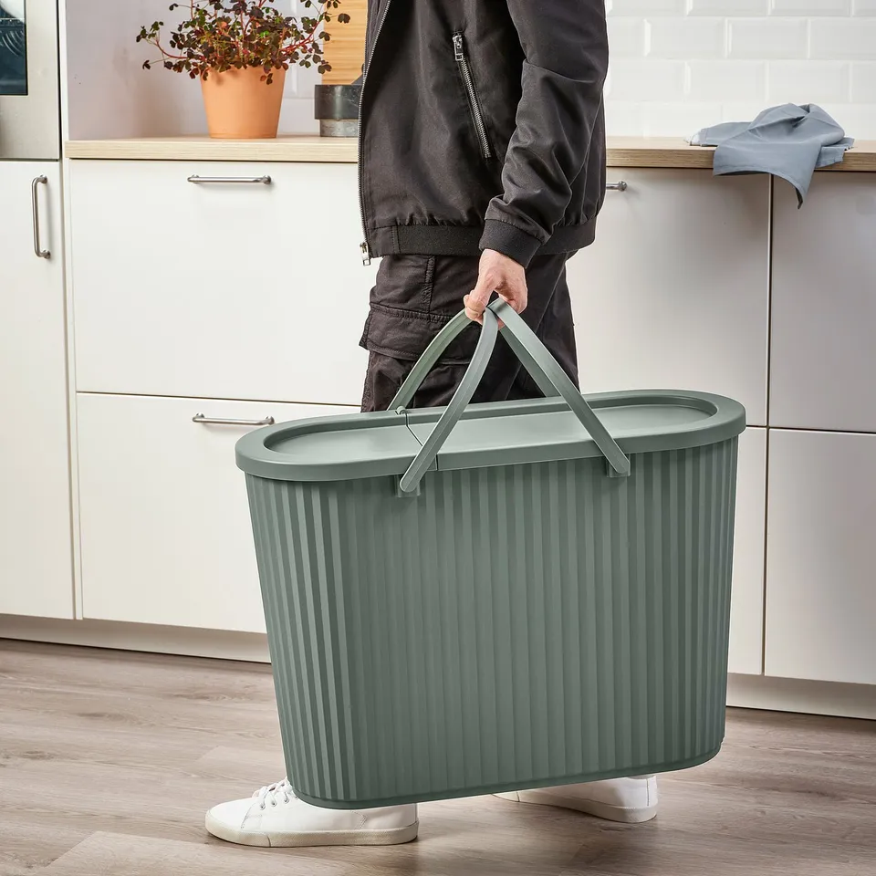 Person carrying green DAMMÄNG bin with lid, suitable for large waste, easy to handle with foldable handle.