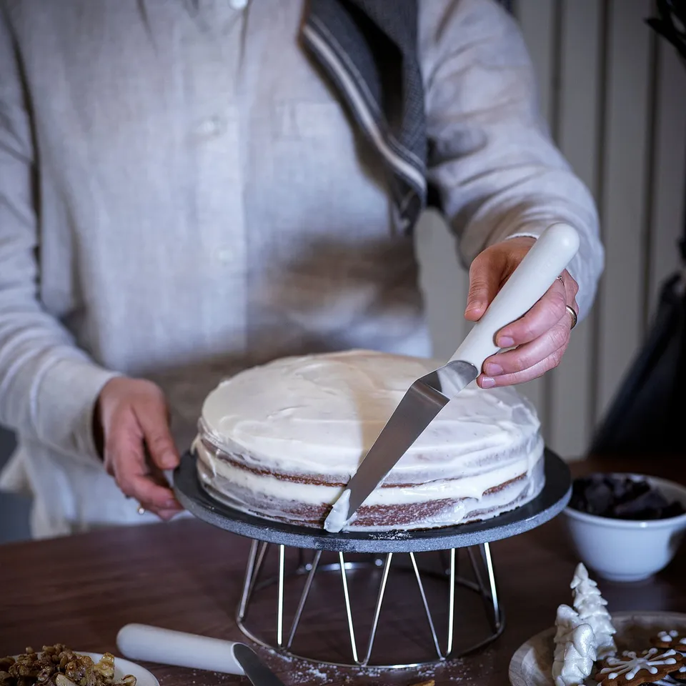 A person cuts a white frosted cake with a white-handled knife on a metal stand. Plates of nuts and cookies surround the cake.