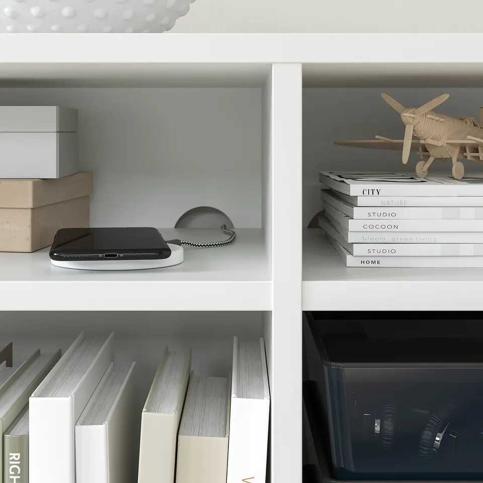 White bookshelf with stacked books, model aeroplane, tech gadget, cardboard boxes, and clear storage bin.