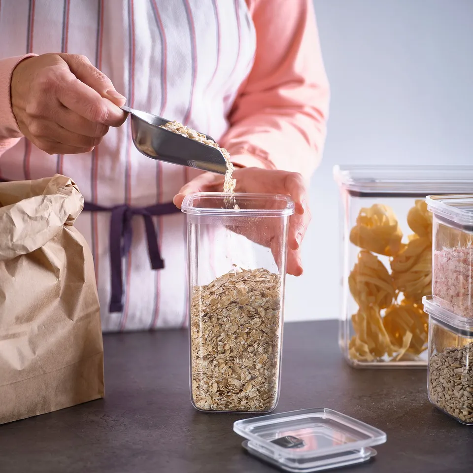 Person pours oats into clear, airtight jar from paper bag. Jars stack and preserve food.