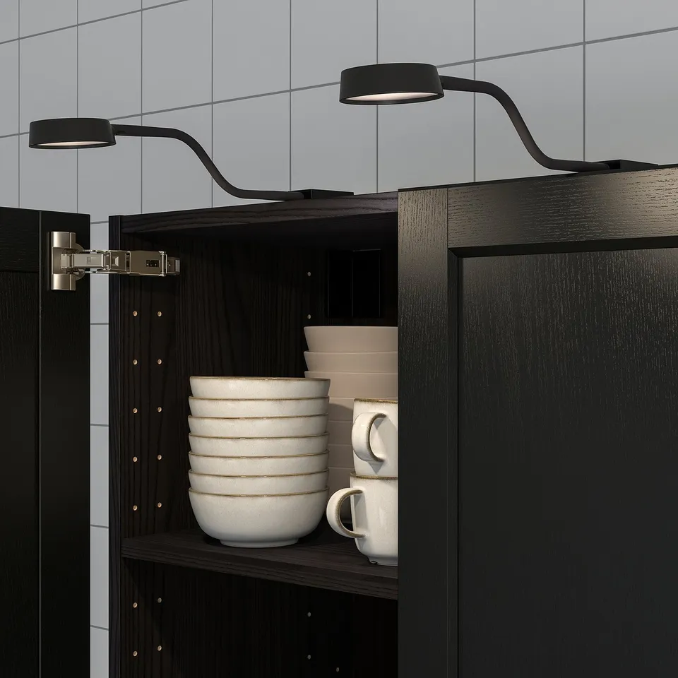 Black kitchen cabinet with stack of bowls and mugs. Two slim, adjustable lights mounted above, illuminating the contents.