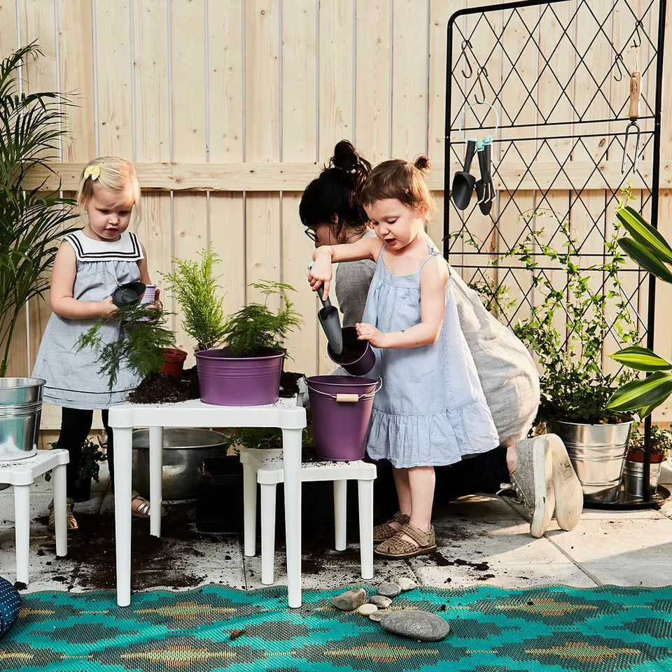 Two young girls in dresses plant herbs on white UTTER childrens tables with purple pots. A green rug and wooden fence background.