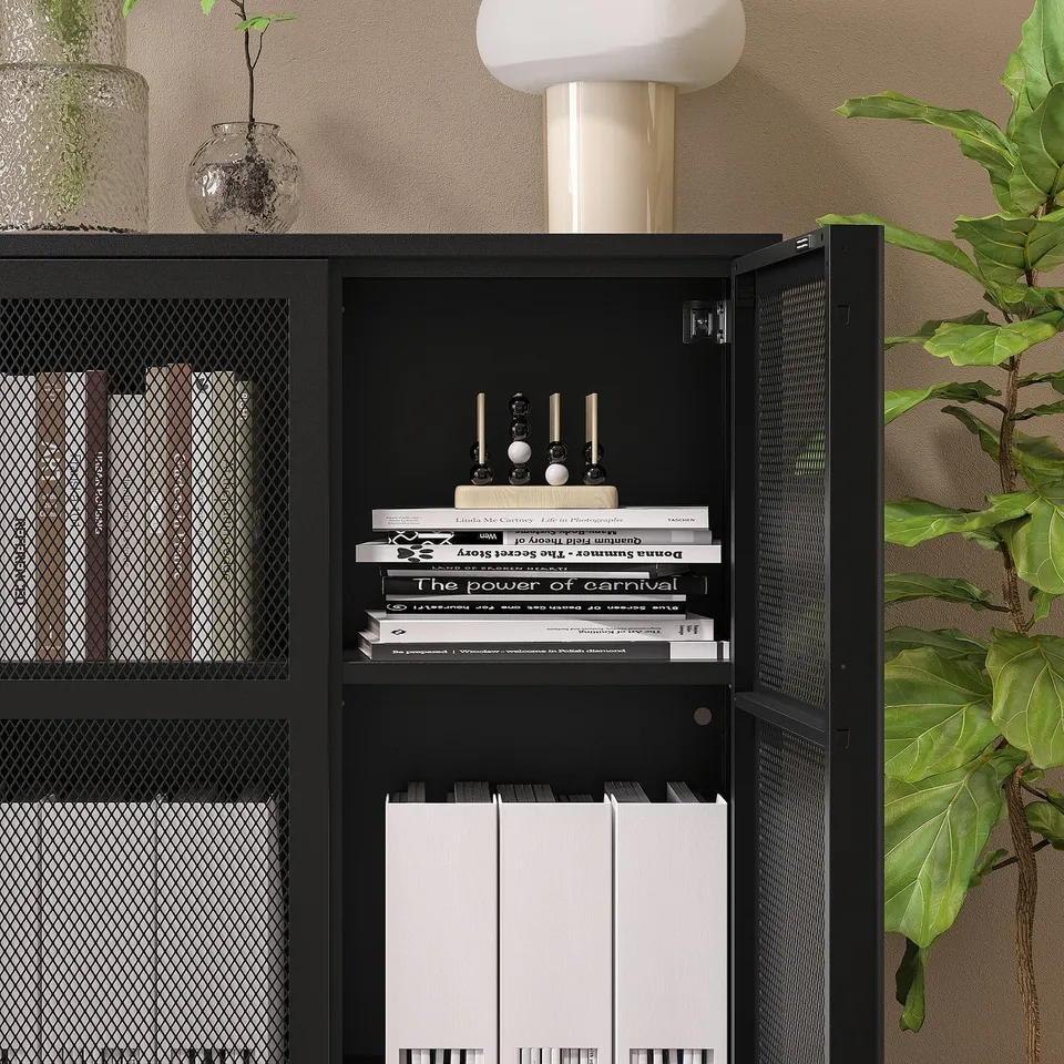 Black IVAR mesh cabinet with adjustable shelves, holding books, and decorative items. A white lamp is on top, with a green plant beside it.