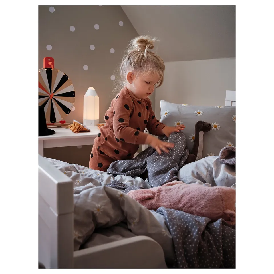 Child playing on bed with NATTSLÄNDA daisy pillow, light pink polka dot pyjamas, soft bedding, night light, and decorative toys.