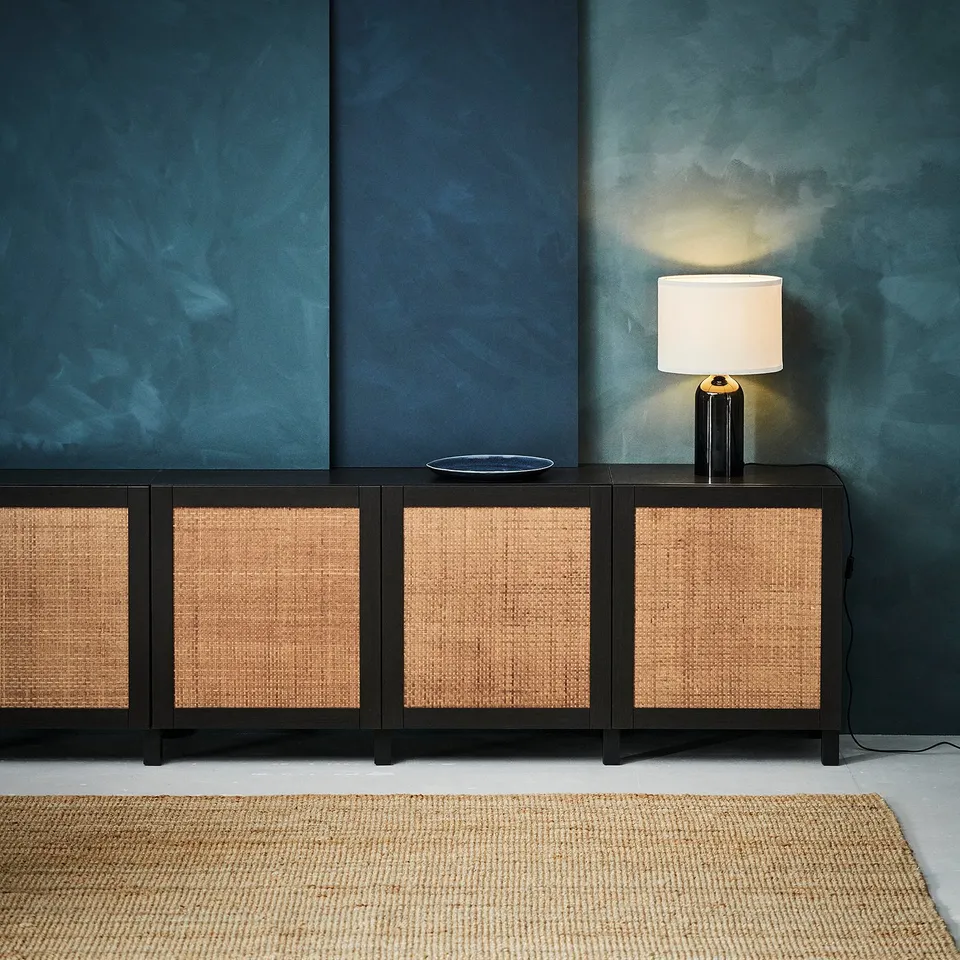 Dark blue wall with black and woven sideboard, lit lamp, beige rug.