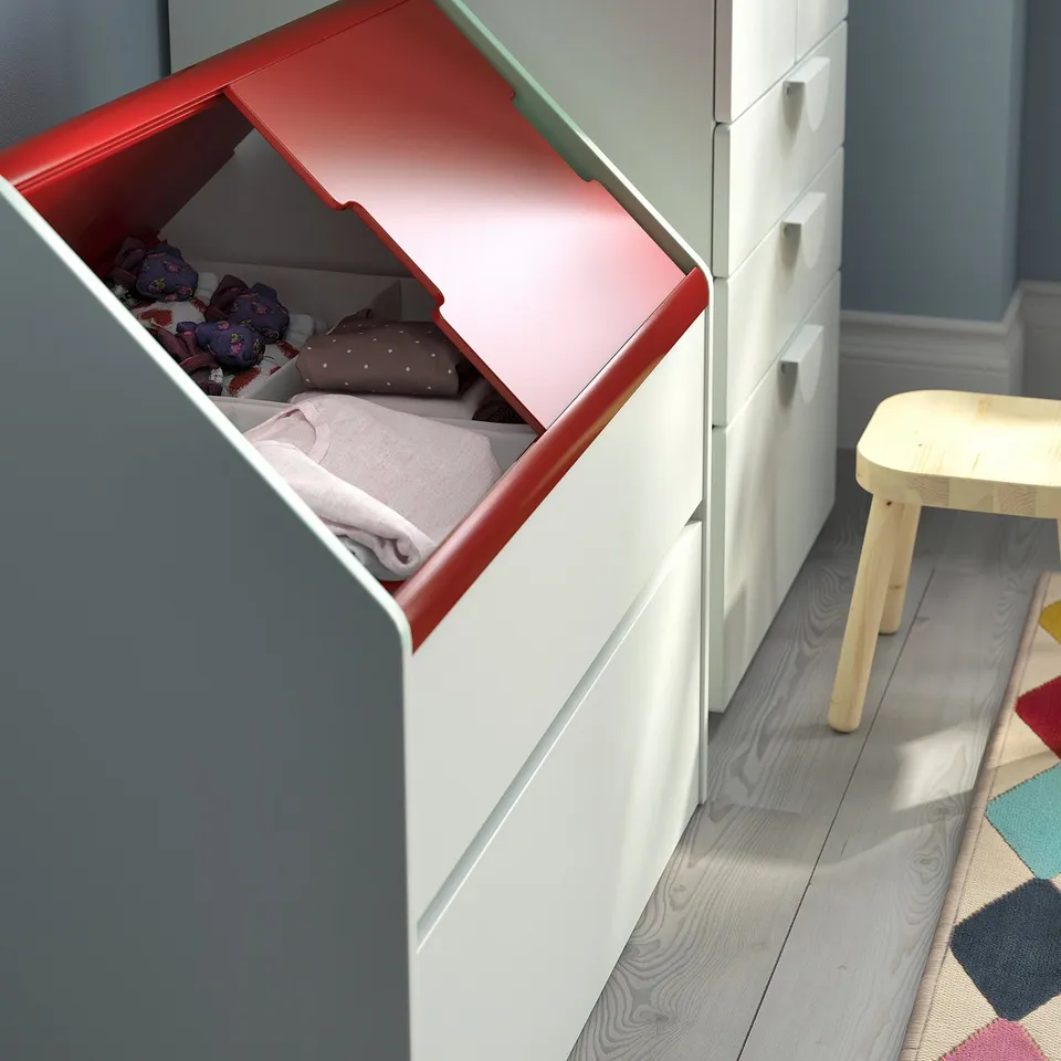 Open red storage box with blankets inside, next to white drawers and a wooden stool on a patterned rug.