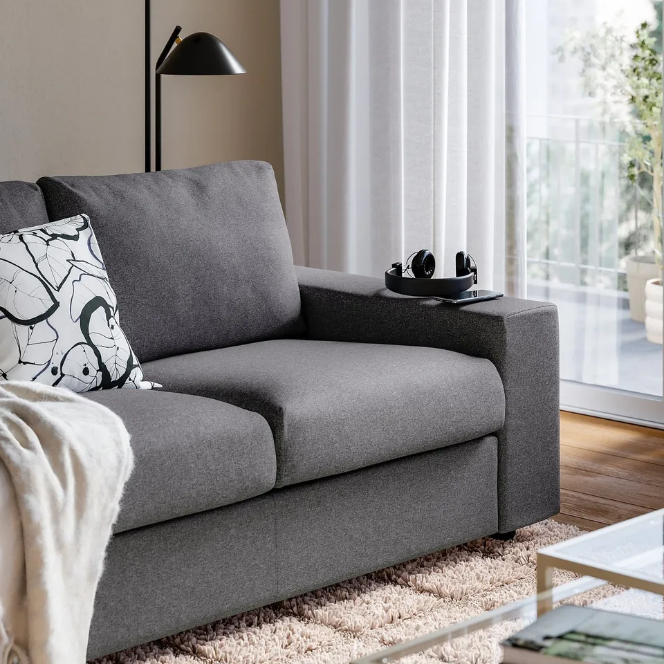 Grey VIMLE sofa with armrests holding headphones, near a window with white curtains and a black lamp.