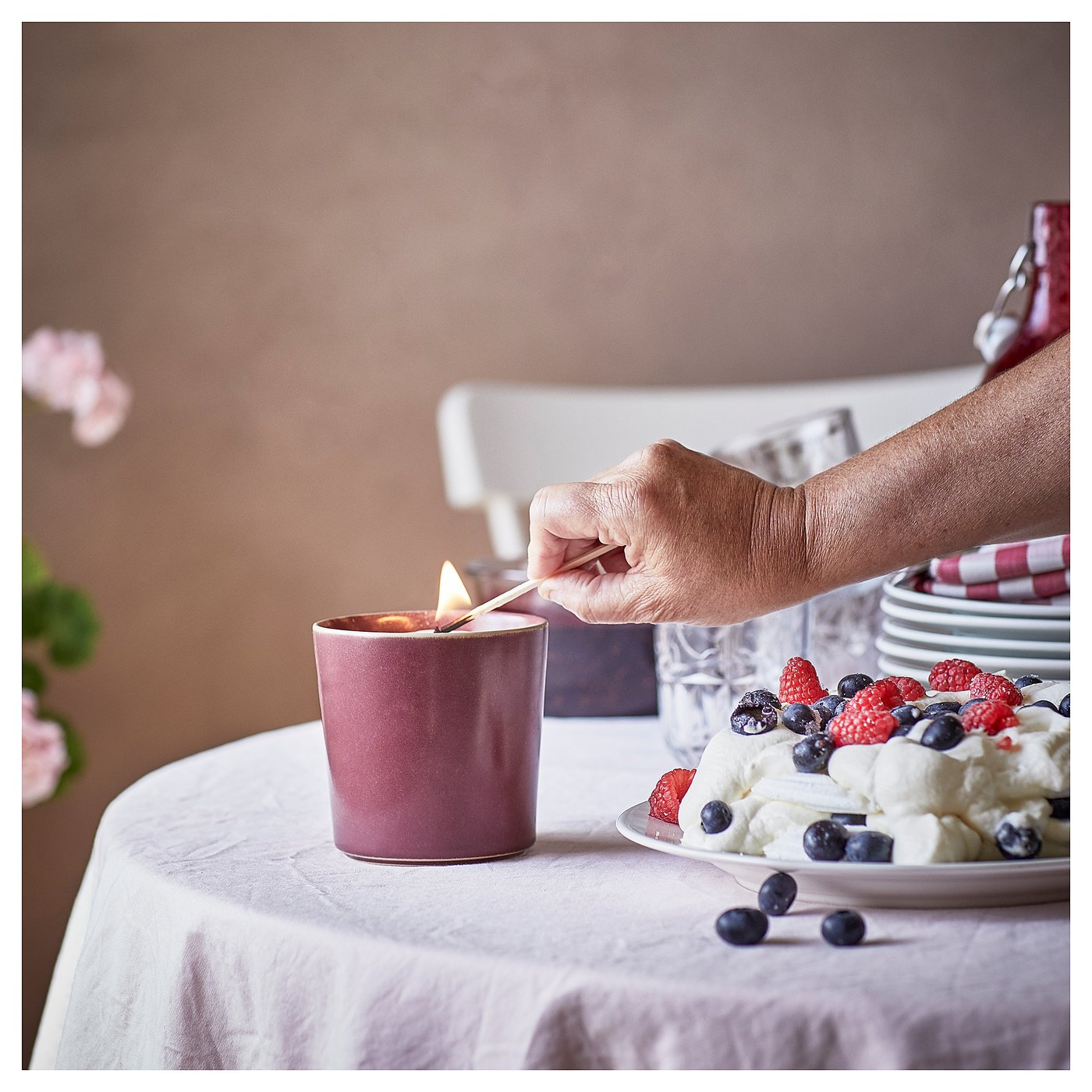 Person lighting pinkish STÖRTSKÖN candle beside fresh fruits and desserts on table.