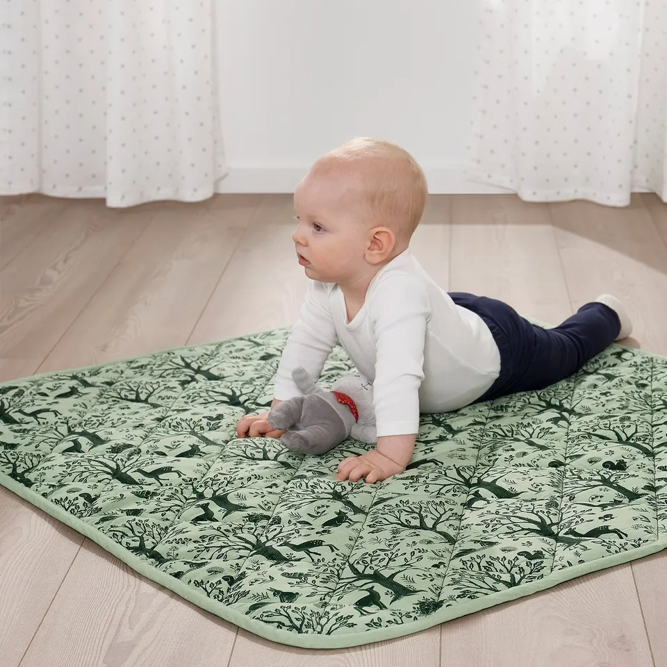 Baby lies on TROLLDOM quilted blanket with forest patterns on a wooden floor. The blanket supports and comforts the infant during playtime.