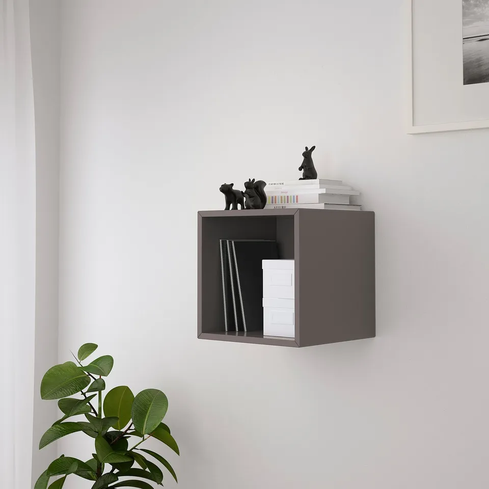 A wall-mounted cube shelf, grey EKET, holding books and small sculptures. Its securely fixed to the wall, offering storage and organisation.