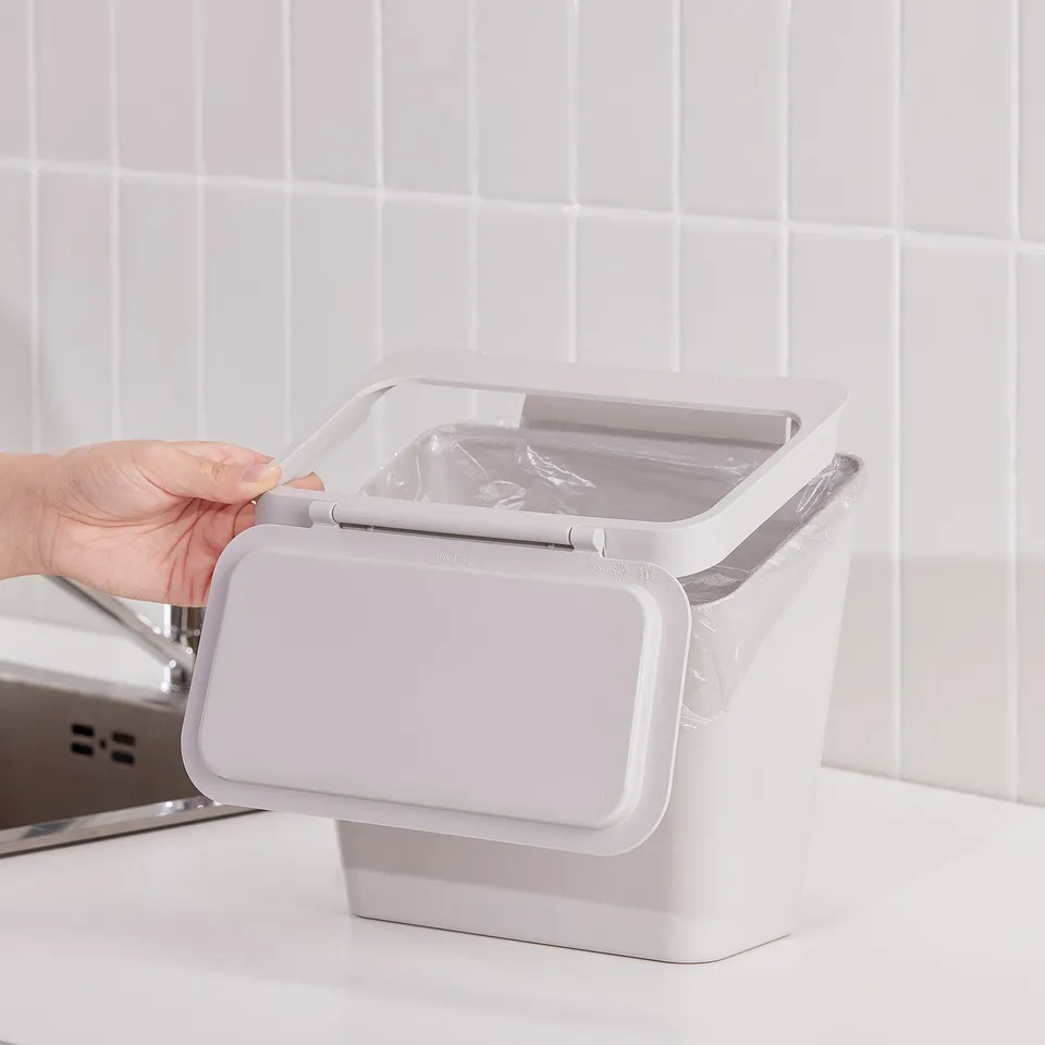 Person opening SKOLÄST bin, a compact waste solution that hangs over doors, ideal for kitchen use.