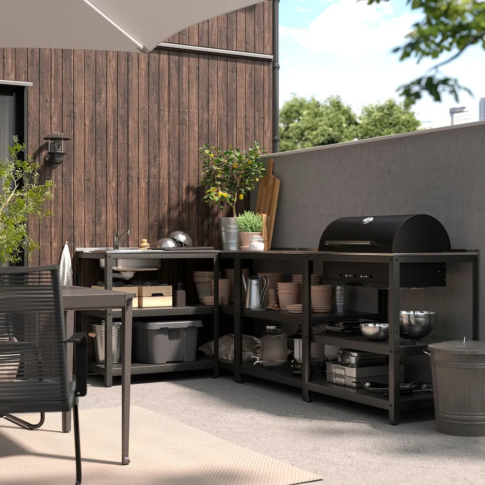 Outdoor kitchen setup with GRILLSKÄR: black grill and shelves, wood panel wall, potted plant, metal sink, seating area.