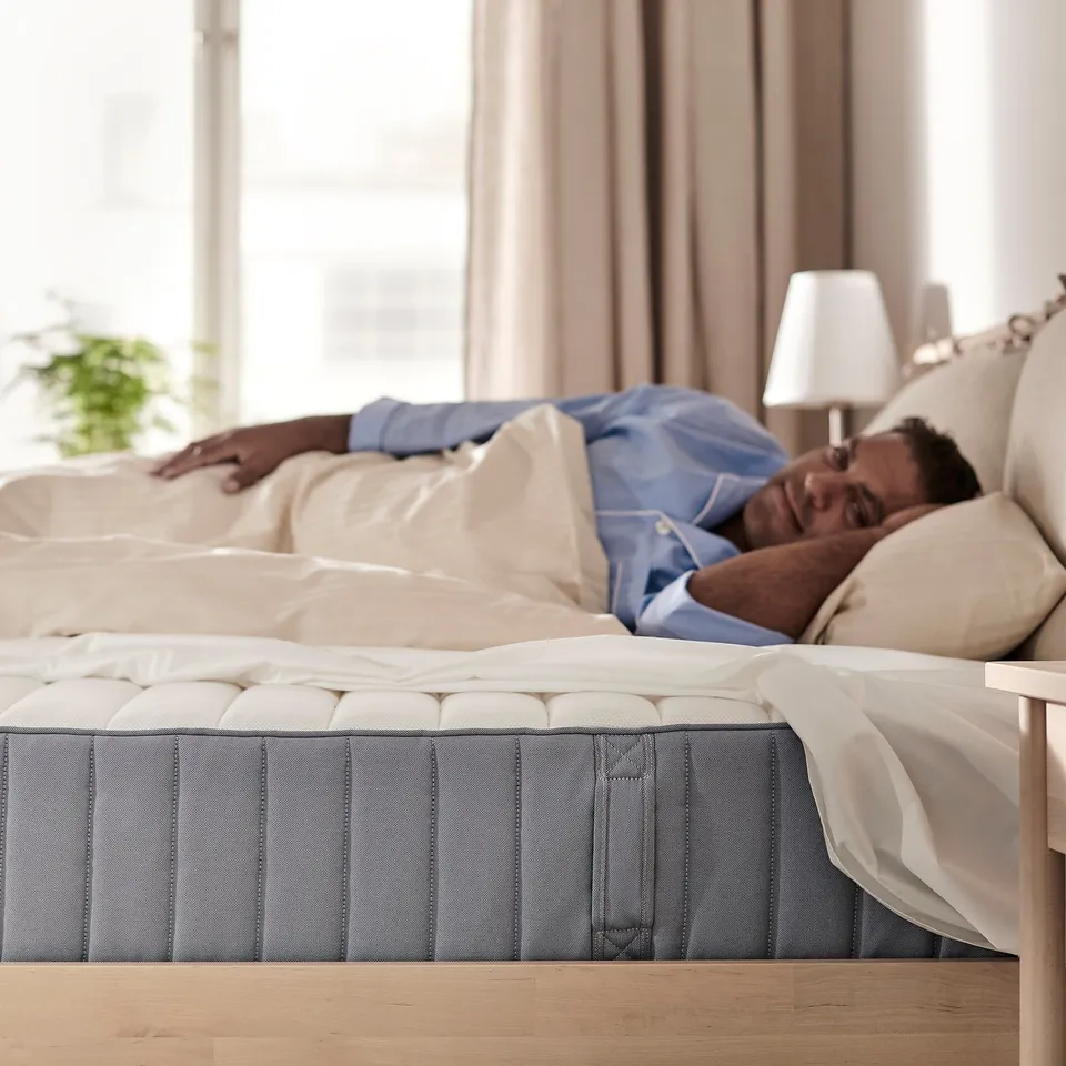 Person sleeping on a firm mattress. Bed is against a window with beige curtains. Mattress has individual pocket springs for support.