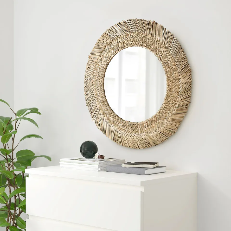 Round mirror with woven frame above cabinet, books, sphere, plant.