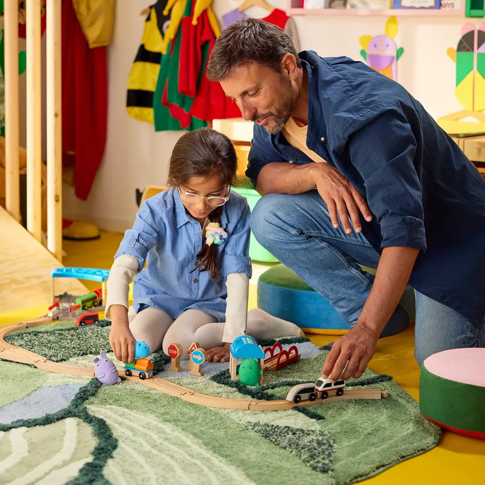 A person and child play with wooden train sets on a large green textured rug, creating a 3d landscape. The rug covers most of the floor space.