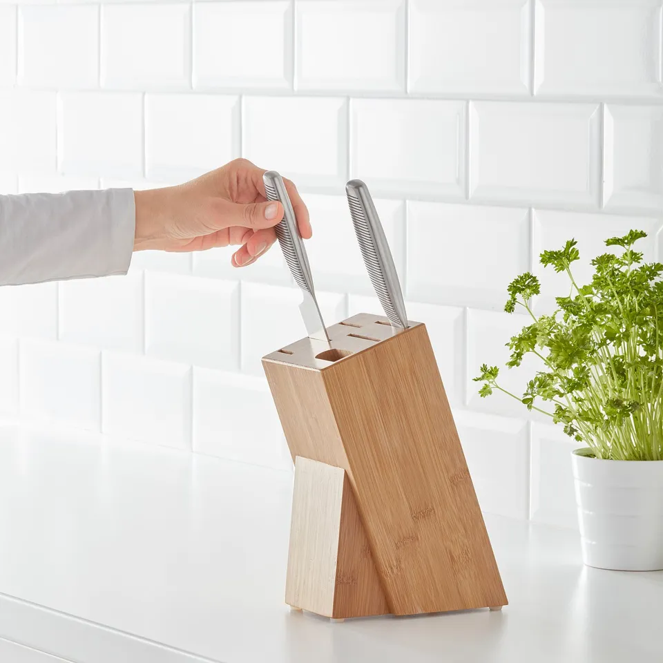 Person grabbing a knife from a wooden HYVLA knife block. Block holds up to 5 knives and sits on a white kitchen counter. A small plant is nearby.