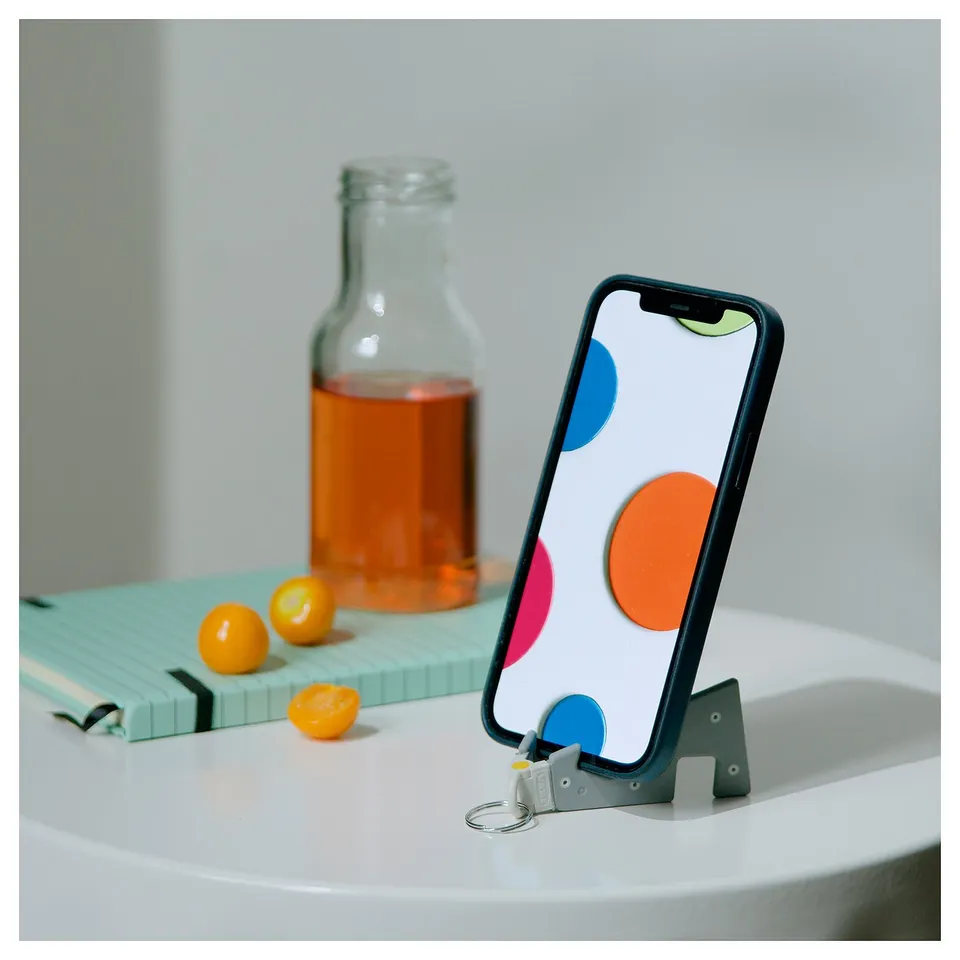 Smartphone holder, black, with colourful dots, stands on table, bottle, citrus fruits, and notepad behind.