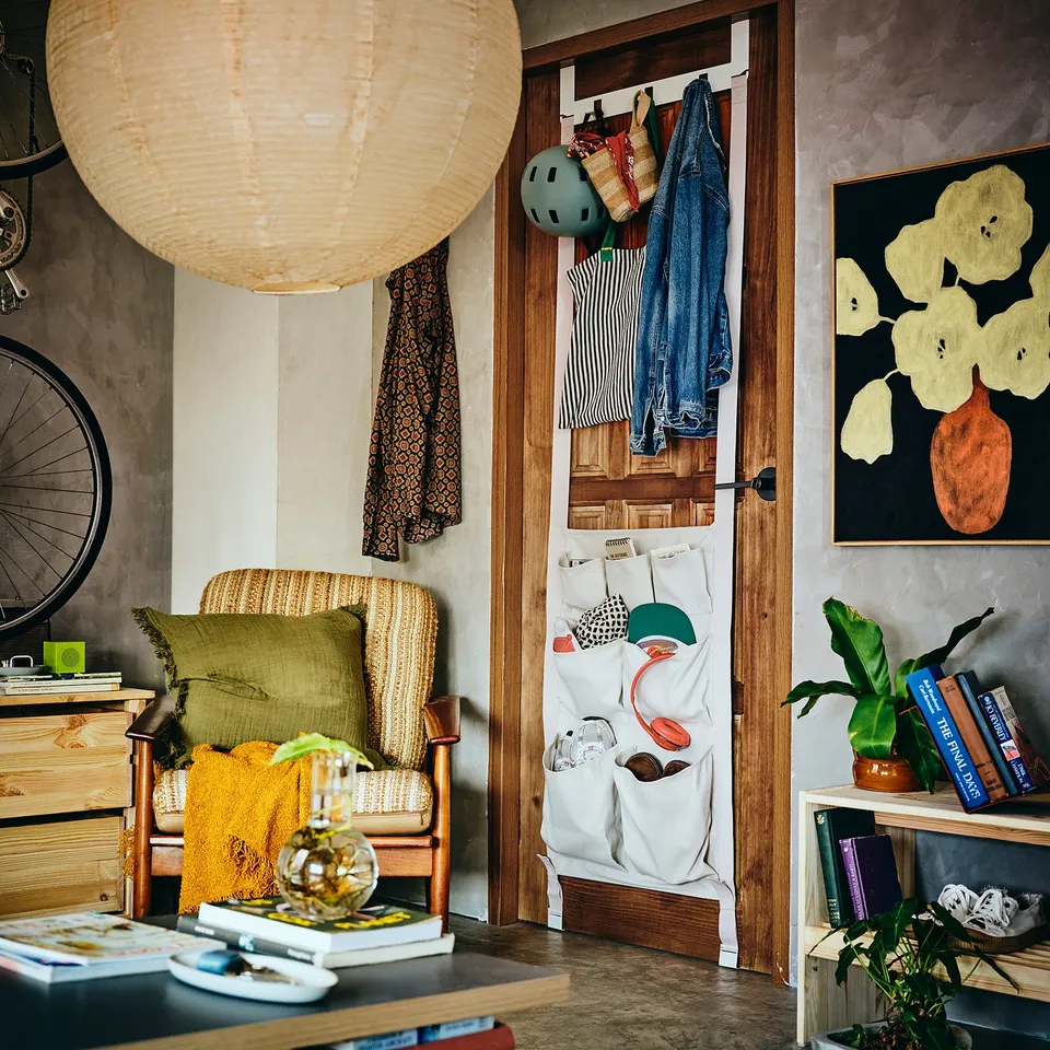 White hanging storage KOMPISHÄNG with pockets and hooks, holding clothes and bags, hanging on a wooden door, surrounded by decor items in a cosy room.