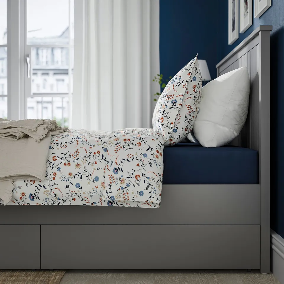 Bed with floral sheets and 2 drawers under frame, large headboard, set against blue wall with large window.