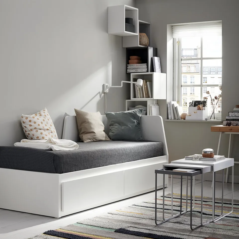 Modern white sofa bed with grey cushions near a large window, accompanied by nesting tables on a striped rug.