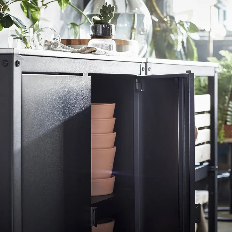 Black outdoor kitchen unit with stacked terracotta pots inside, green plants around, stainless steel countertop with decorative items.