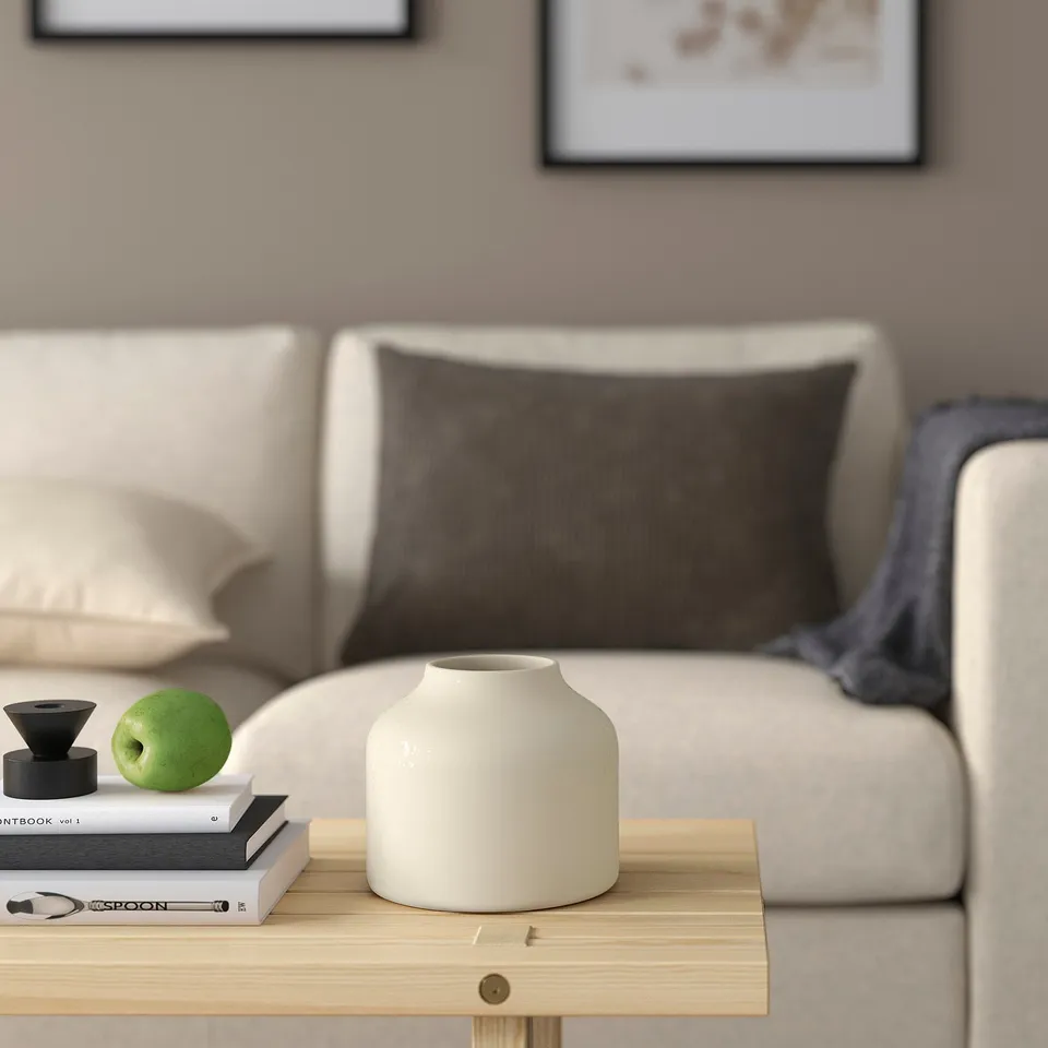 Living room with beige sofa, brown pillow, grey blanket, BLODBJÖRK vase on wooden table with books and apple.