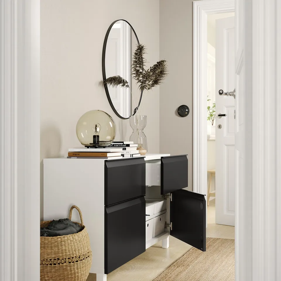 Modern entrance with white and black BESTÅ sideboard, round mirror, plant, lamp, books, and basket.