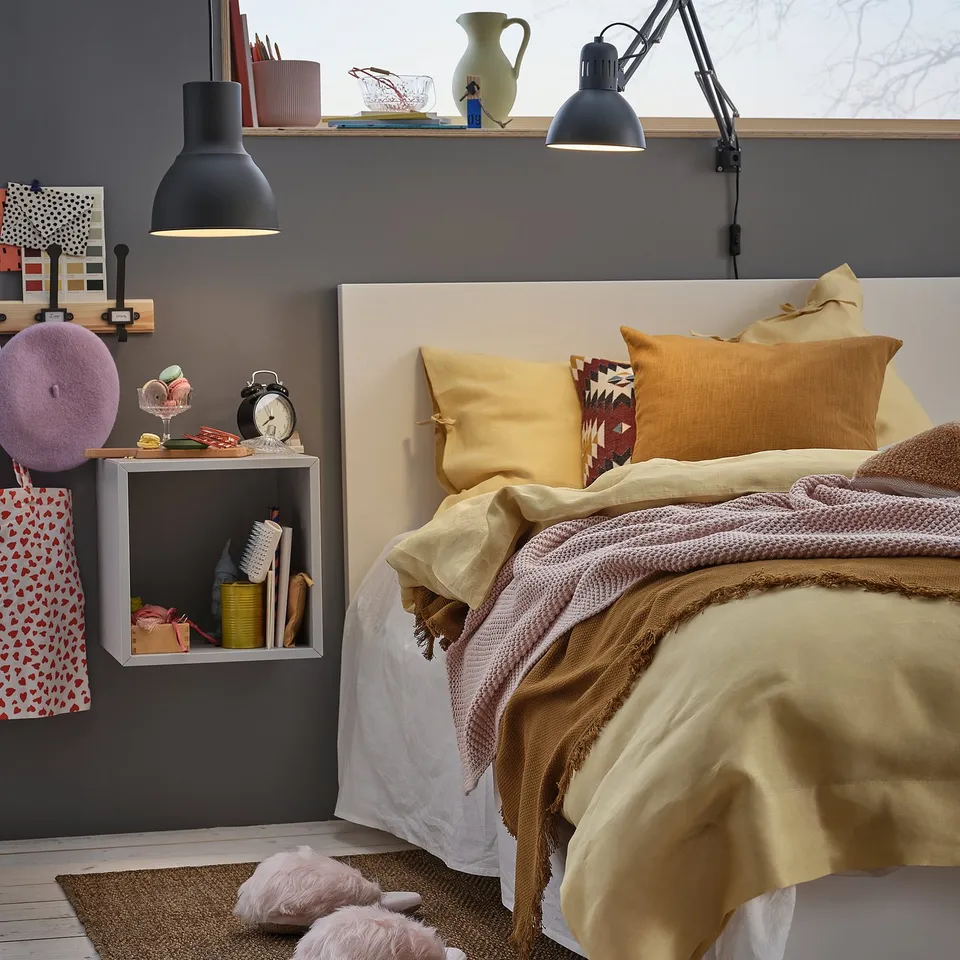 Cosy bedroom with beige bedding, colourful pillows, and a TERTIAL black adjustable lamp. Decorated shelves, rugs, and slippers.