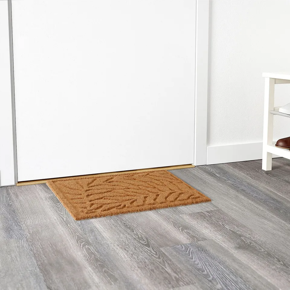 Brown 3d leaf-patterned doormat on grey wooden floor, against white door and bench.