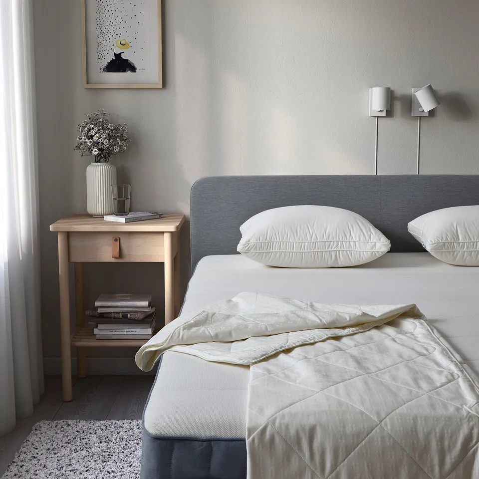 Twin bed with white bedding, blue headboard, bedside table, vase, books, candles, wall art, lamps. Bedding retains a cool, cosy look.