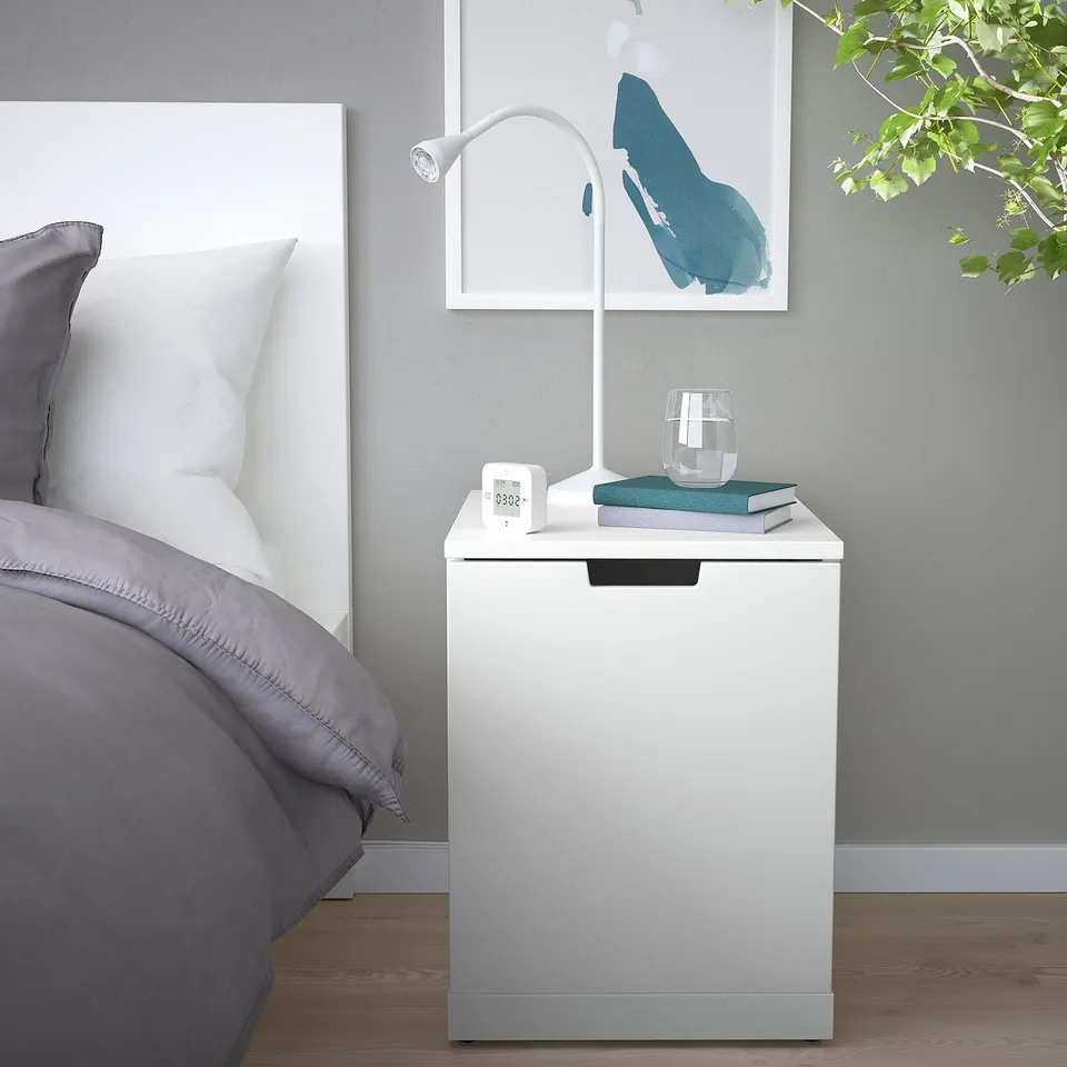 Small white NORDLI bedside table beside bed with grey bedding, holding lamp, books, and glass. Grey wall with hanging art.