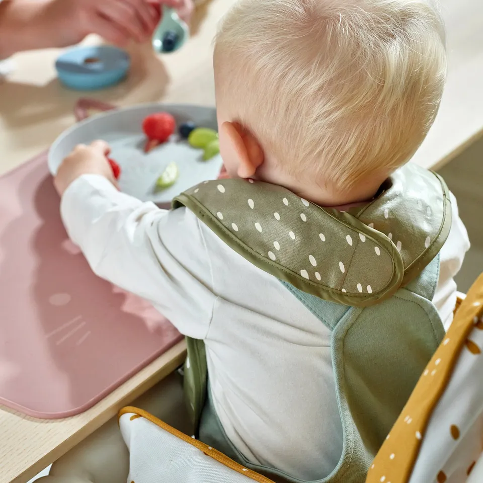 Baby in green bib eats fruit in high chair.