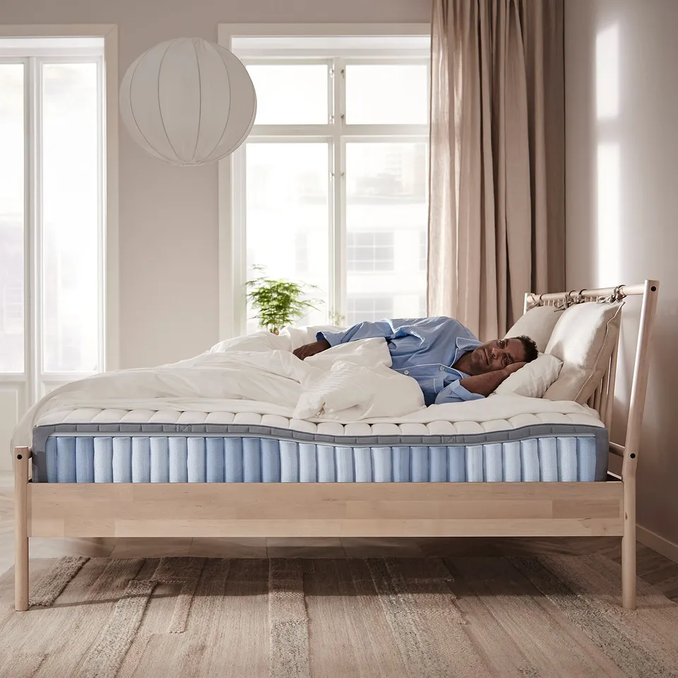 A person sleeps on a light wooden bed with a blue and white mattress. Room has large windows and a hanging white lamp.