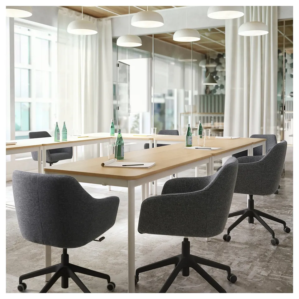 Modern office space with long wooden conference table, grey upholstered chairs, white pendant lights, and natural light from large windows.