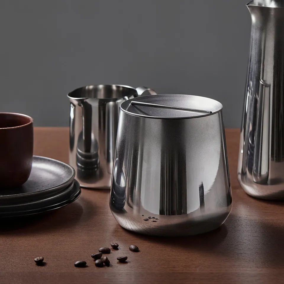 Stainless steel kitchen jar with lid and scattered coffee beans on table. Modern and elegant kitchen storage.