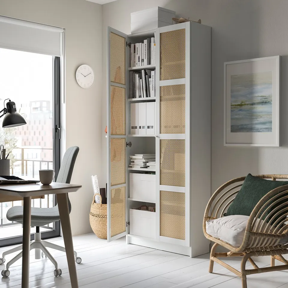 White BILLY bookcase with bamboo doors, containing books, near a window in a minimalist home office.