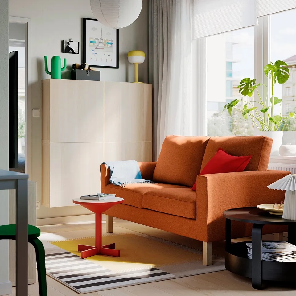 Cosy living room with orange SALTSJÖBADEN sofa, beige cabinet, side table, rug, and plant, large windows.