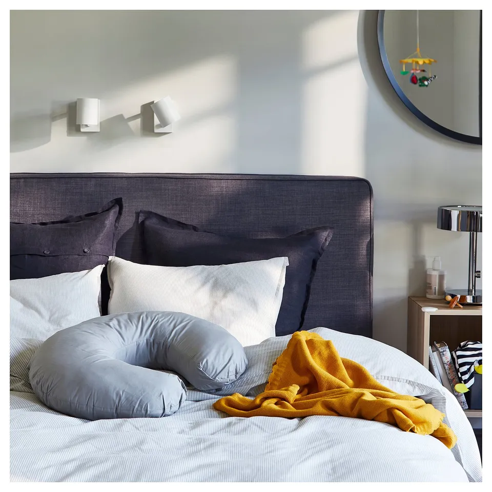 Modern bedroom with grey headboard, pillows, pregnancy pillow, yellow blanket, round mirror, and wooden bedside table with lamp.
