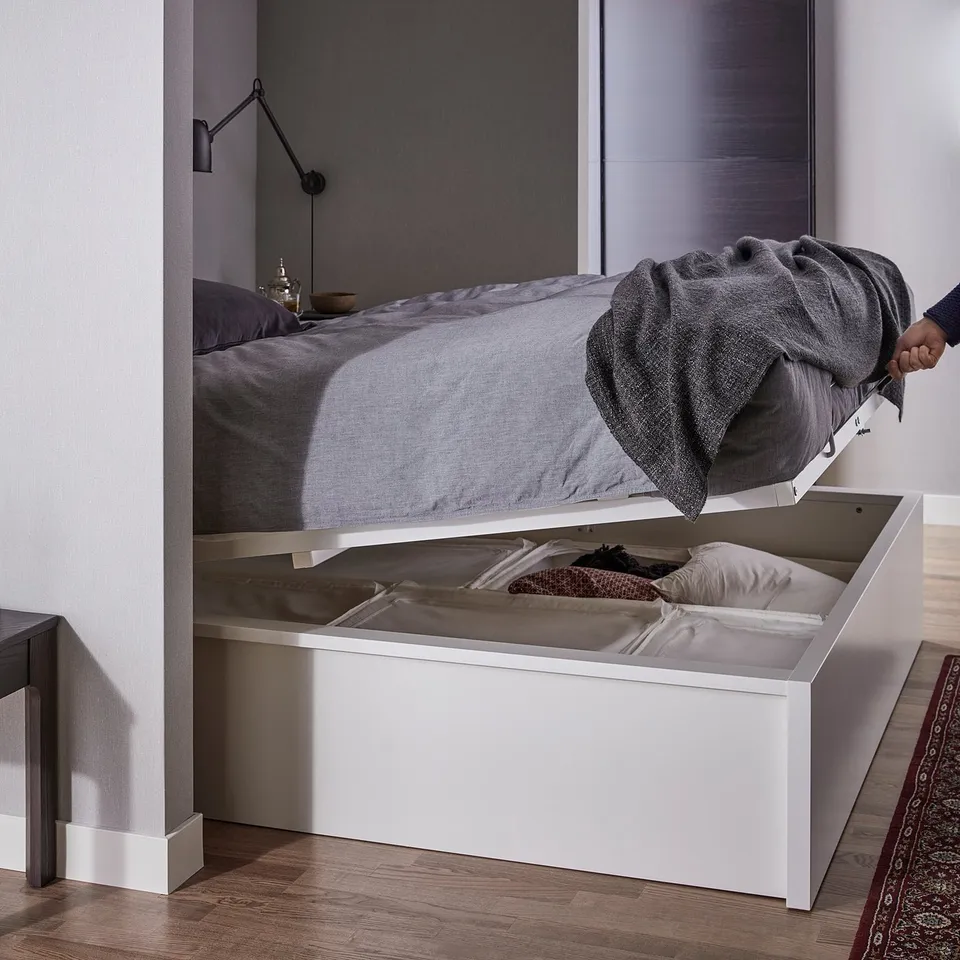 Modern bedroom with white MALM platform bed, grey bedding, and a wall lamp. A human hand is opening the storage compartment beneath the bed.