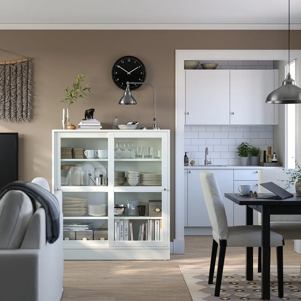 Modern living room with HAVSTA shelf, dishes, decor, clock, lamp, rug. Kitchen with white cabinets, grey chairs, plants.