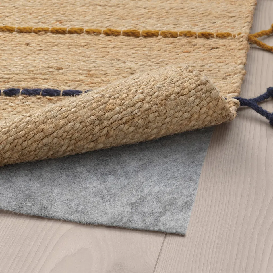 Natural jute rug with wool border sits on grey felt mat on wooden floor, showing rugs edge and underside.