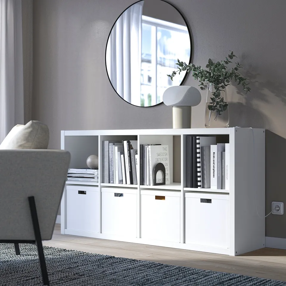 KALLAX white bookshelf with various compartments and decor items like books and plants. Mirror and chair nearby.