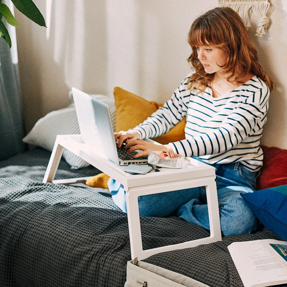 A person works on a laptop with a KLIPSK tray on the person bed, book, backpack, and trainers nearby.