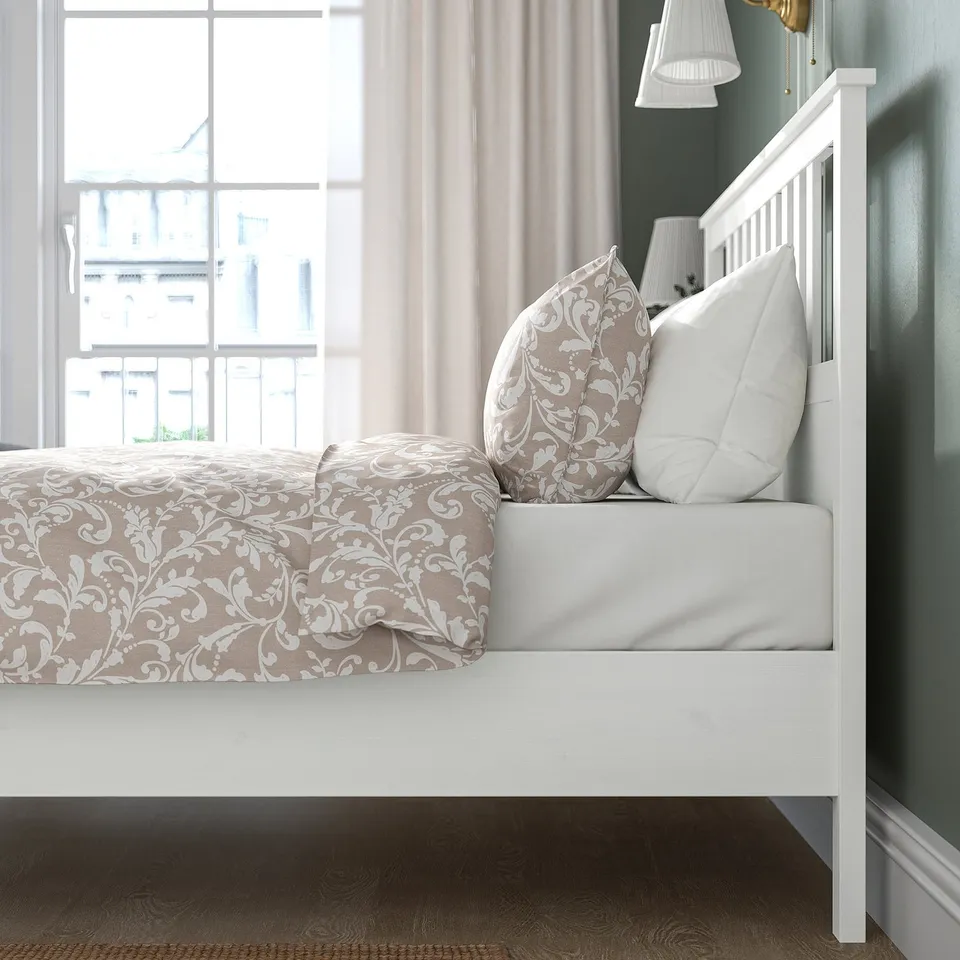 A white wooden bed with intricate bedding sits by a window, showcasing its high headboard and footboard for comfort and stability.