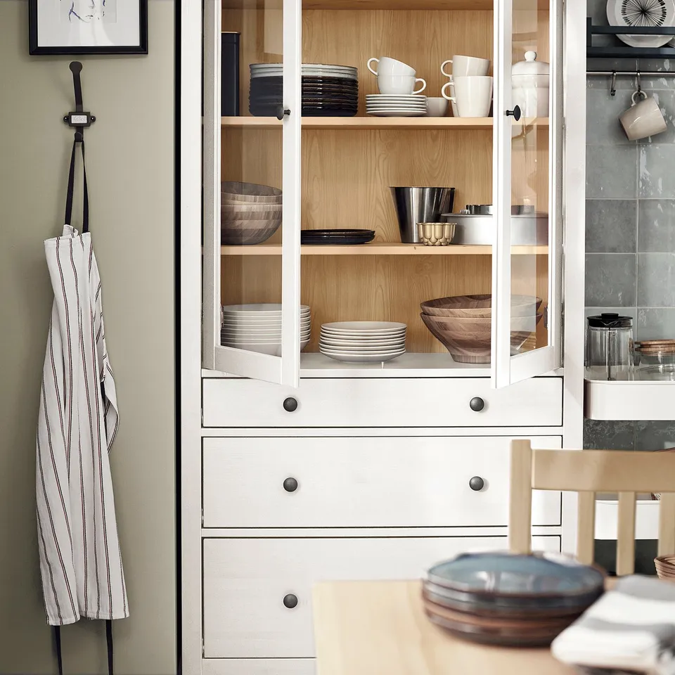 HEMNES white wooden cabinet, with glass doors, holds stacked dishes, wooden bowls, and cups. An apron hangs beside it, emphasising its size and style.