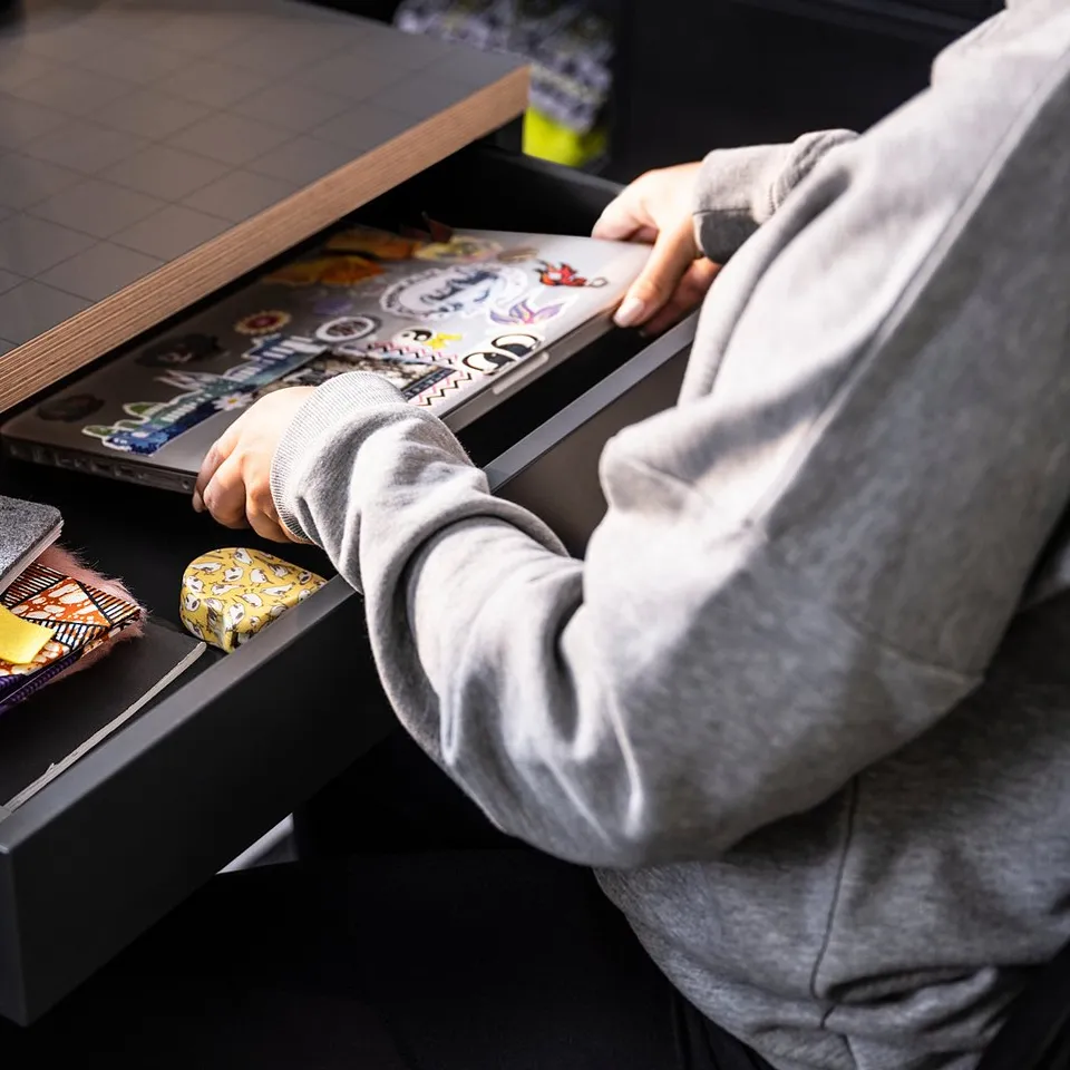 Person in grey hoodie reaching into black MICKE desk drawer with colourful notebooks, a yellow object, and cables.
