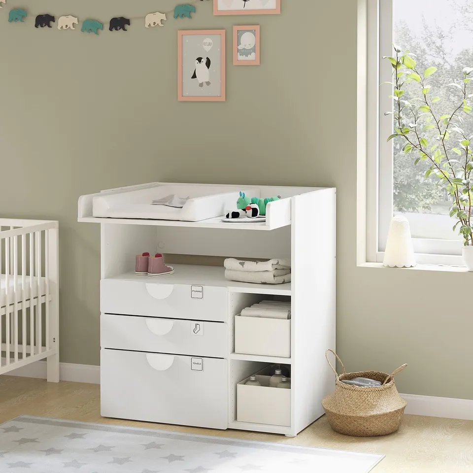 White nursery changing table with drawers and shelves, baby items, green walls, wooden floor.
