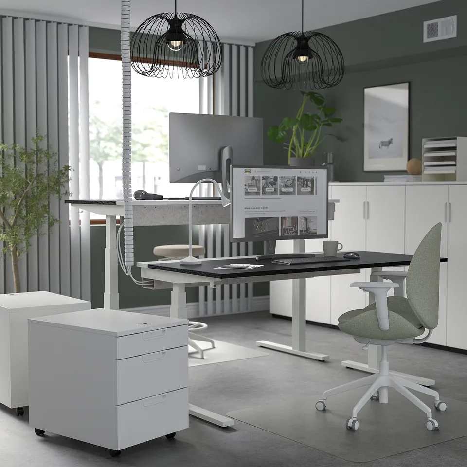 Modern green office with MITTZON desk, ergonomic chair, & storage. Desk has monitor, keyboard, plants, & 2 black pendant lights.