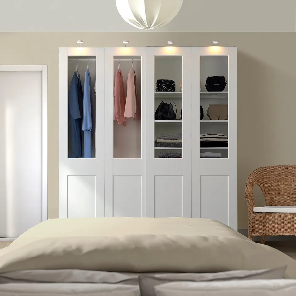 Modern bedroom with large white PAX wardrobe featuring glass doors, displaying clothes, and a beige bed.