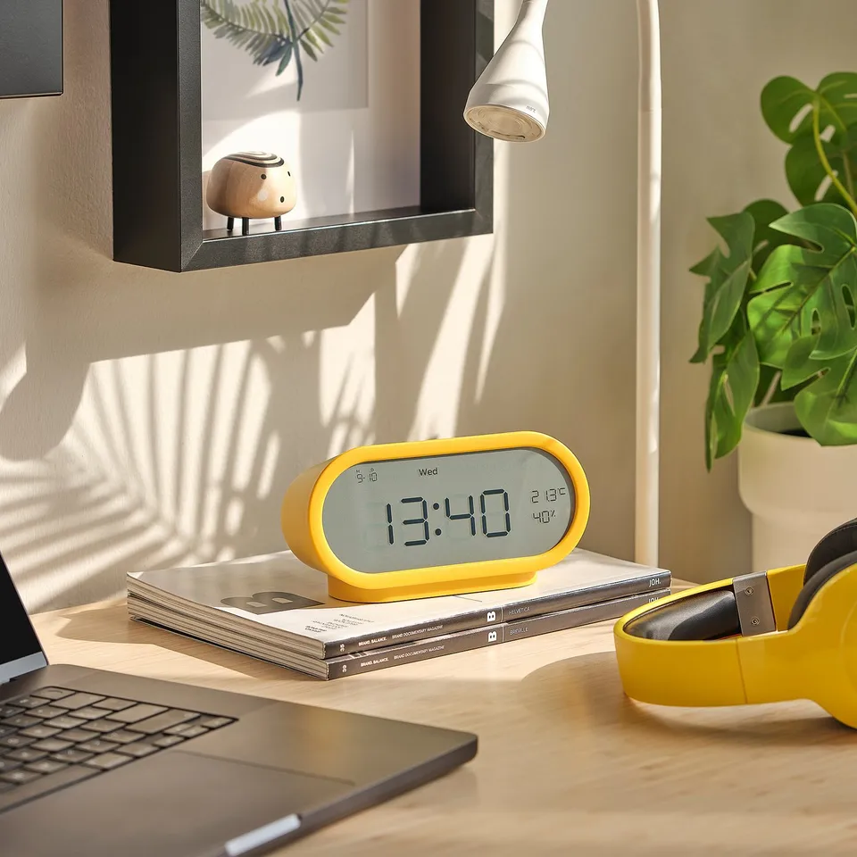 Yellow RÖDSTRÄFSE alarm clock on desk with time, date, and temperature, near laptop and headphones, in natural light.